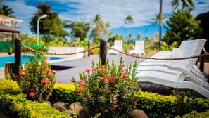 Un espace aménagé au bord de la piscine, avec des chaises longues, une barrière en corde et des arbustes en fleurs.