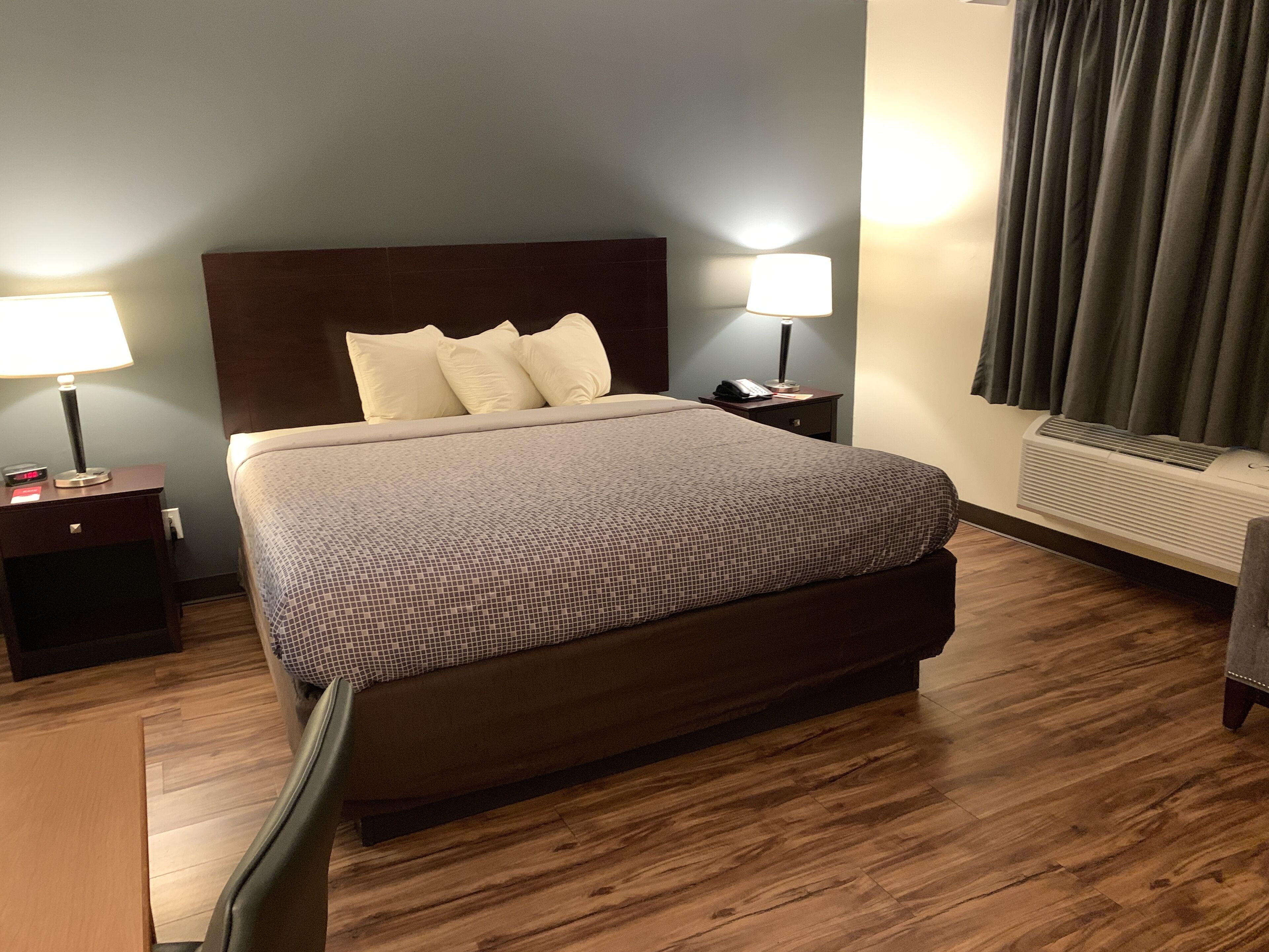 A hotel room with a large bed, two bedside lamps, a nightstand, a heater, and a window with curtains.