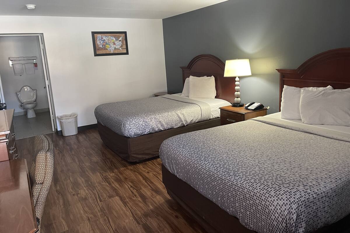 Habitación de hotel con dos camas, suelo de madera, baño con inodoro y lavabo, y una mesita con lámpara.