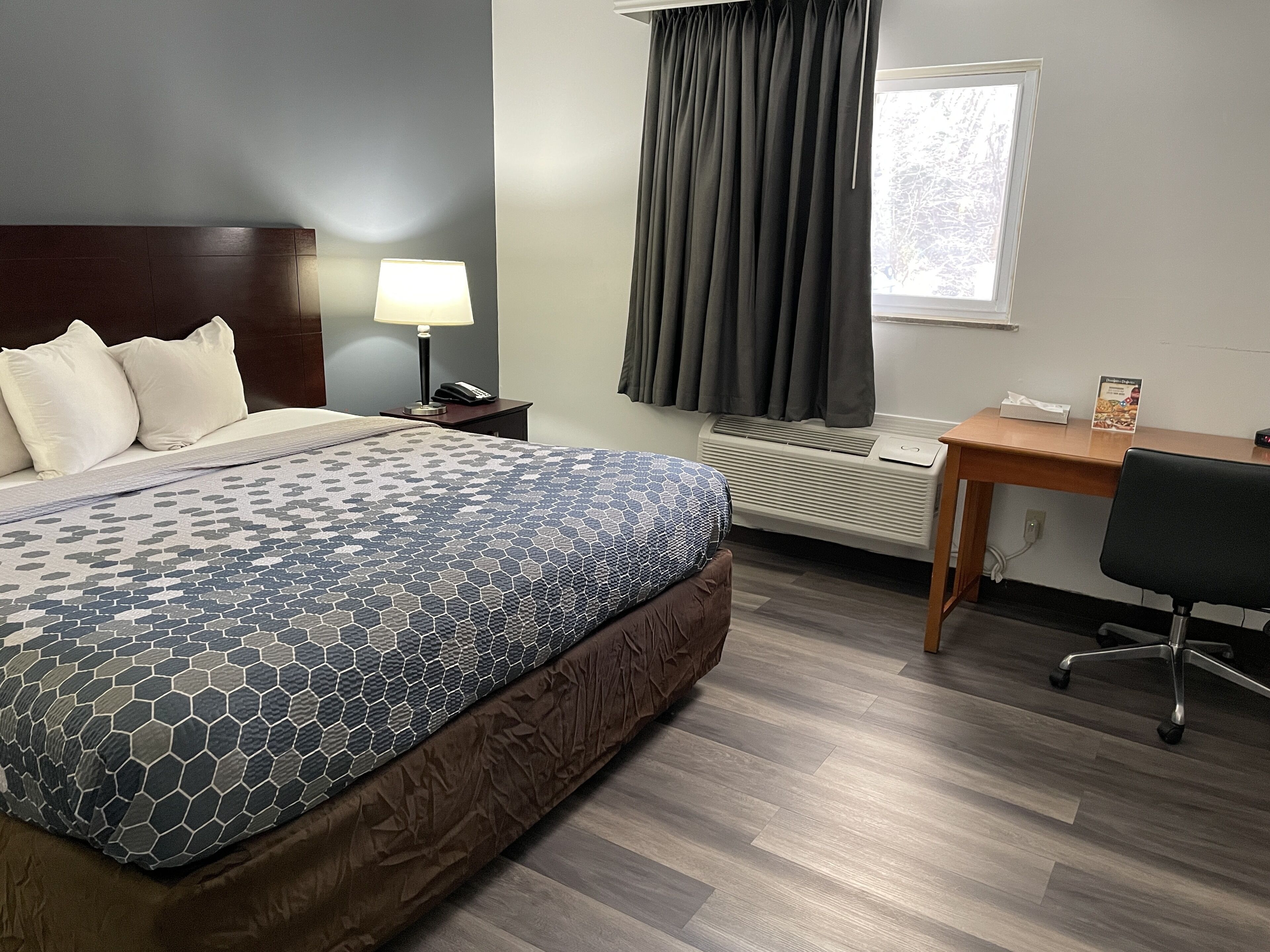 A hotel room with a bed, a desk, a chair, a lamp, a telephone, a window with curtains, and an air conditioning unit.
