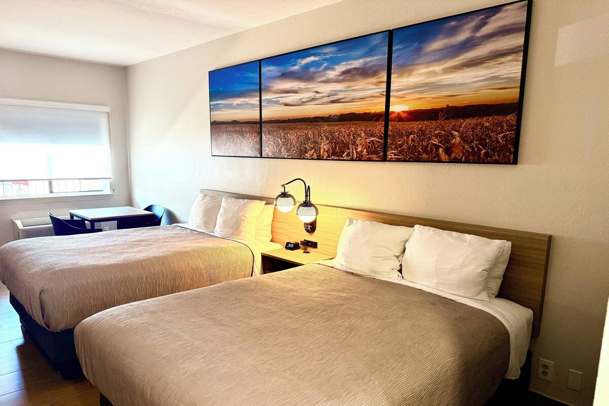Une chambre d’hôtel avec deux lits, une table de chevet avec une lampe, une fenêtre donnant sur un champ et un grand tableau représentant un paysage accroché au mur.