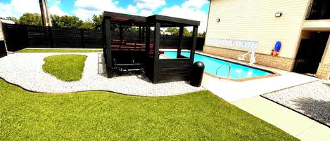 A backyard with artificial grass, a pool, a wooden pergola, and a building with a brick facade.