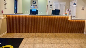 A hotel reception desk with a 'Quality by Choice Hotels' sign, a digital welcome screen, and a reception area with a tiled floor.