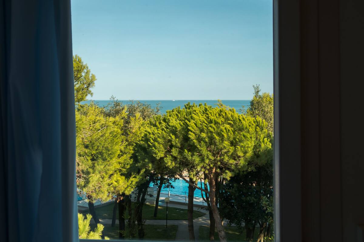 Blick auf einen von Bäumen umgebenen Pool, mit blauem Himmel und weiter Wasserfläche im Hintergrund.