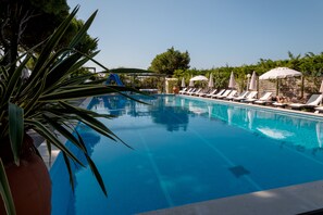 Piscina con sillones de descanso y sombrillas, rodeada de vegetación.
