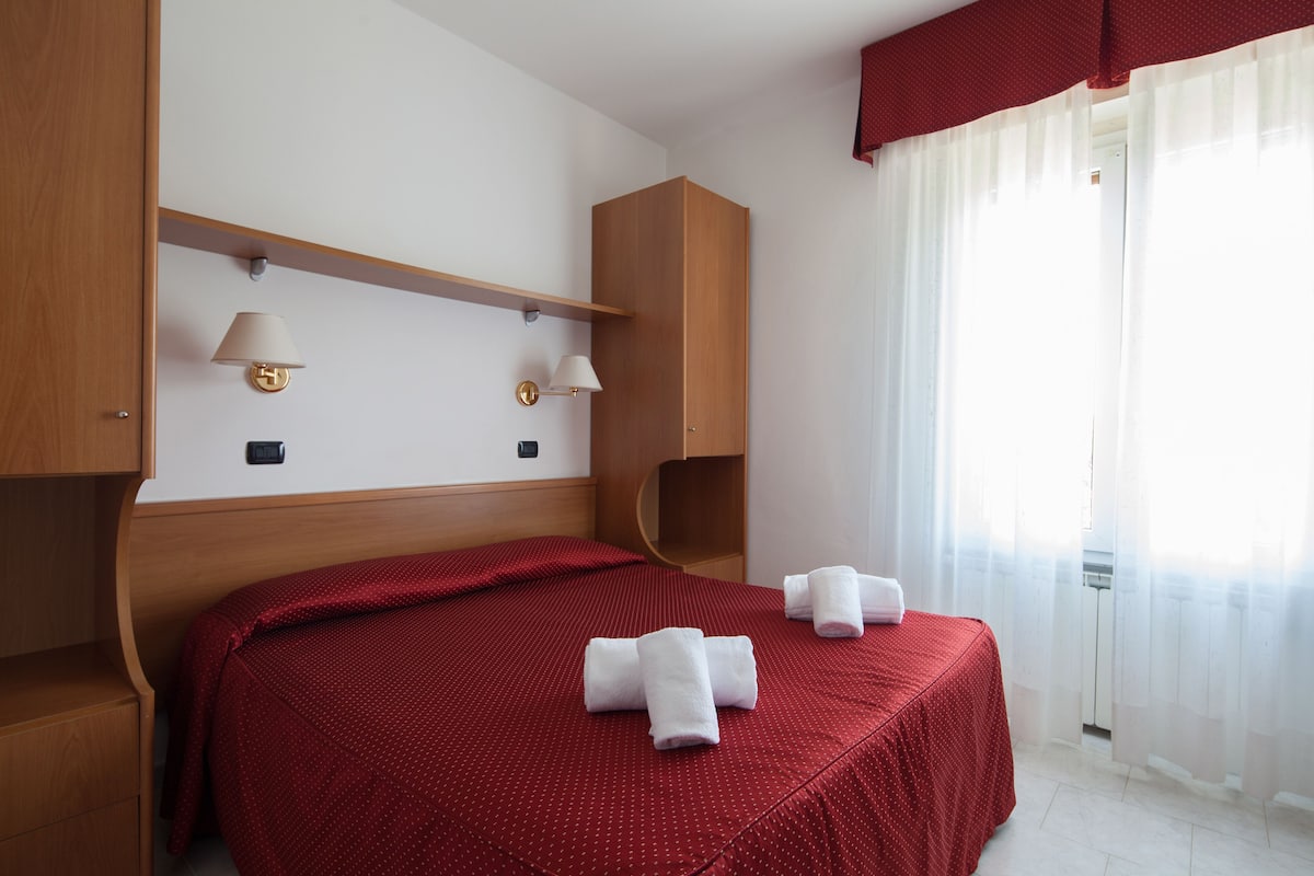 Habitación de hotel con cabecera de madera, colcha roja, dos lámparas fijadas a la pared y cortina roja.