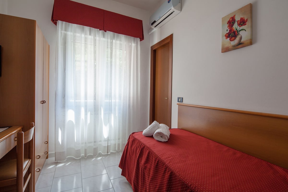 Habitación de hotel con cama, escritorio, silla, ventana con cortinas translúcidas y unidad de aire acondicionado.
