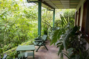 A covered outdoor area with green chairs and a table, surrounded by lush greenery.