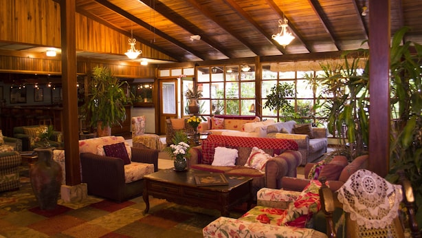 A cozy, well-lit lounge area with wooden beams, comfortable seating, and large windows offering a view of greenery outside.