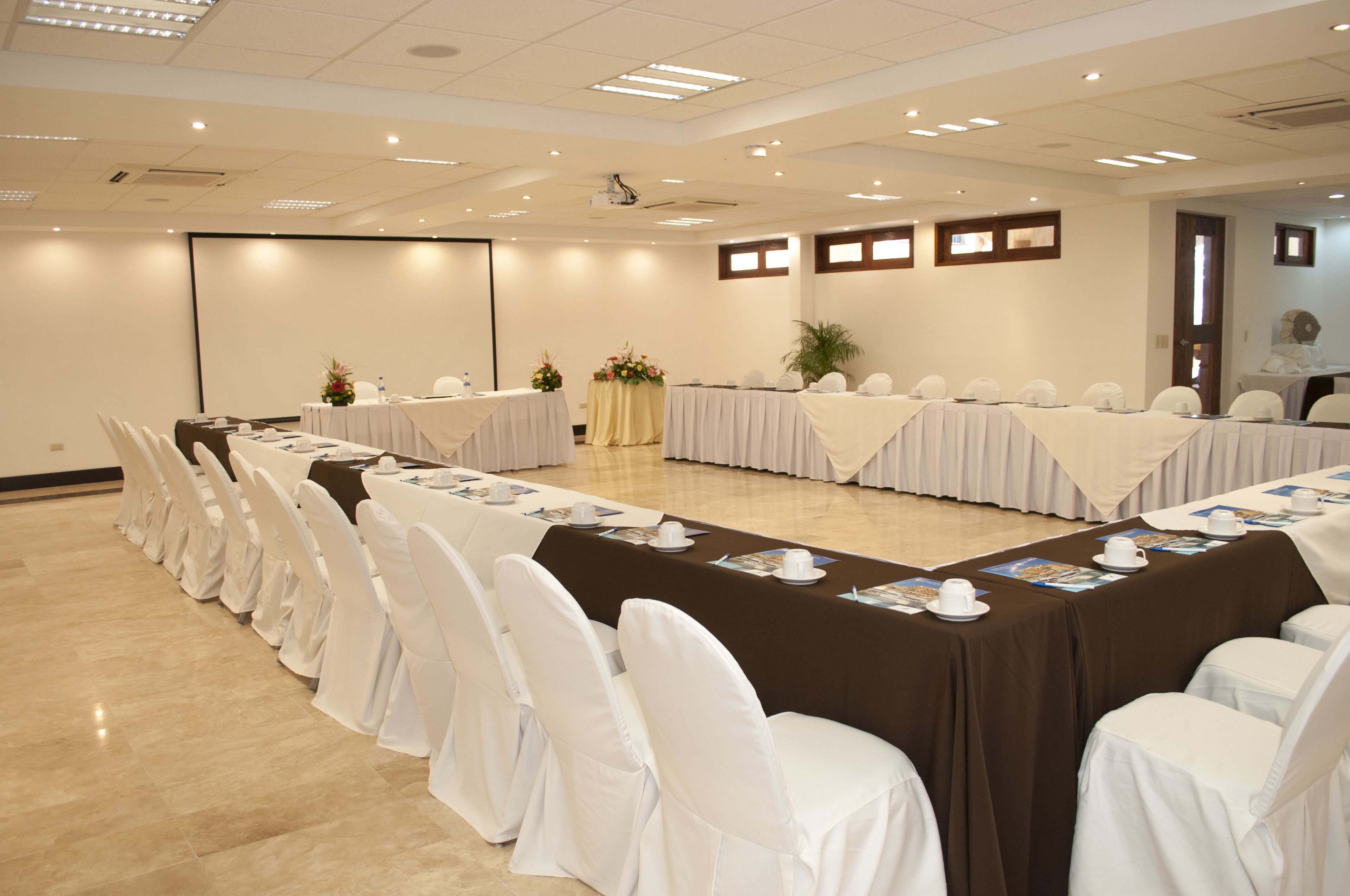 Un salón de conferencias con largas mesas, sillas blancas y una pantalla de proyector.