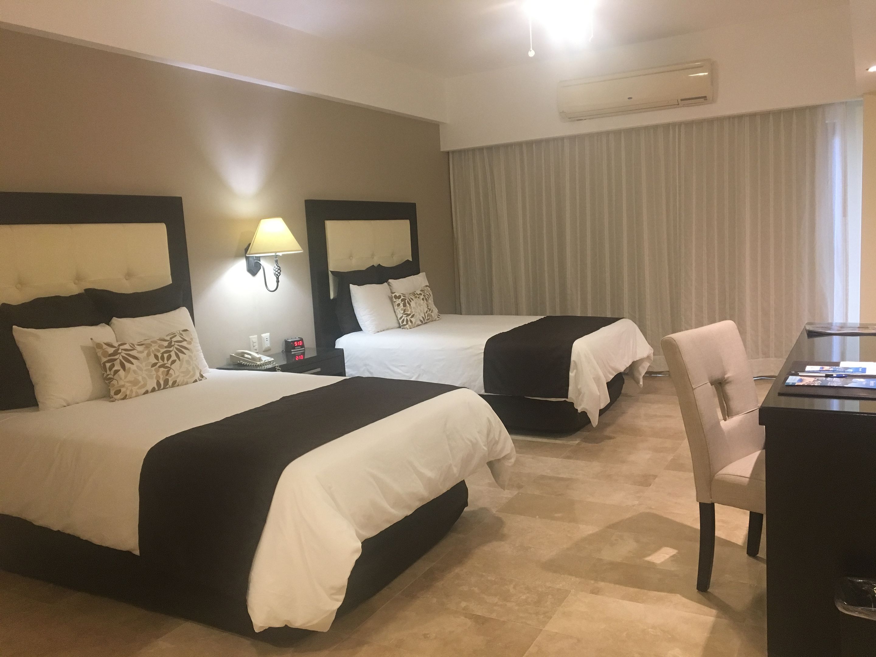 A hotel room with two beds, a desk, a chair, a lamp, and an air conditioning unit.