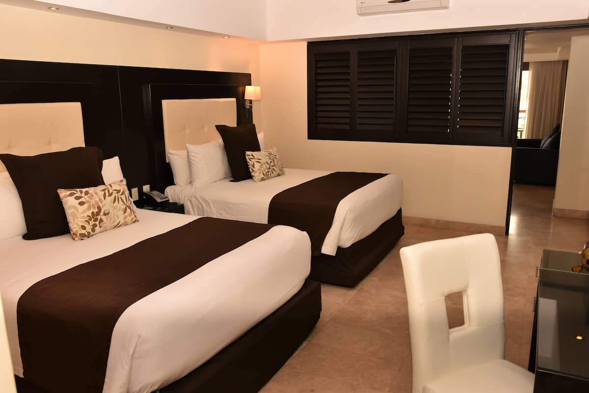 Habitación de hotel con dos camas, cada una con cabecera, un escritorio con silla y una ventana con persianas.