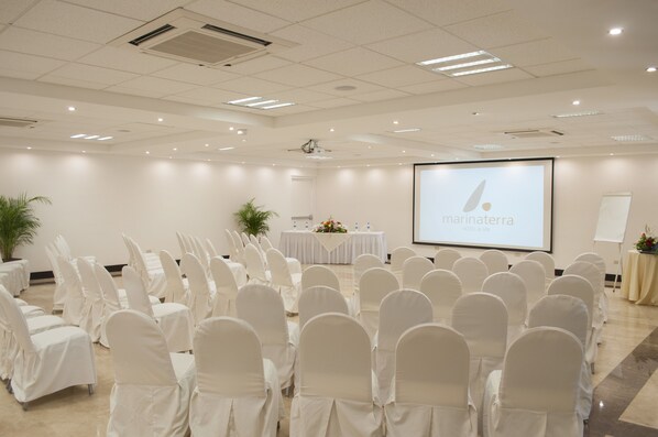 Un salón de conferencias con hileras de sillas blancas, una pantalla mostrando 'marin terra hotel & spa' y un escenario con una mesa y flores.