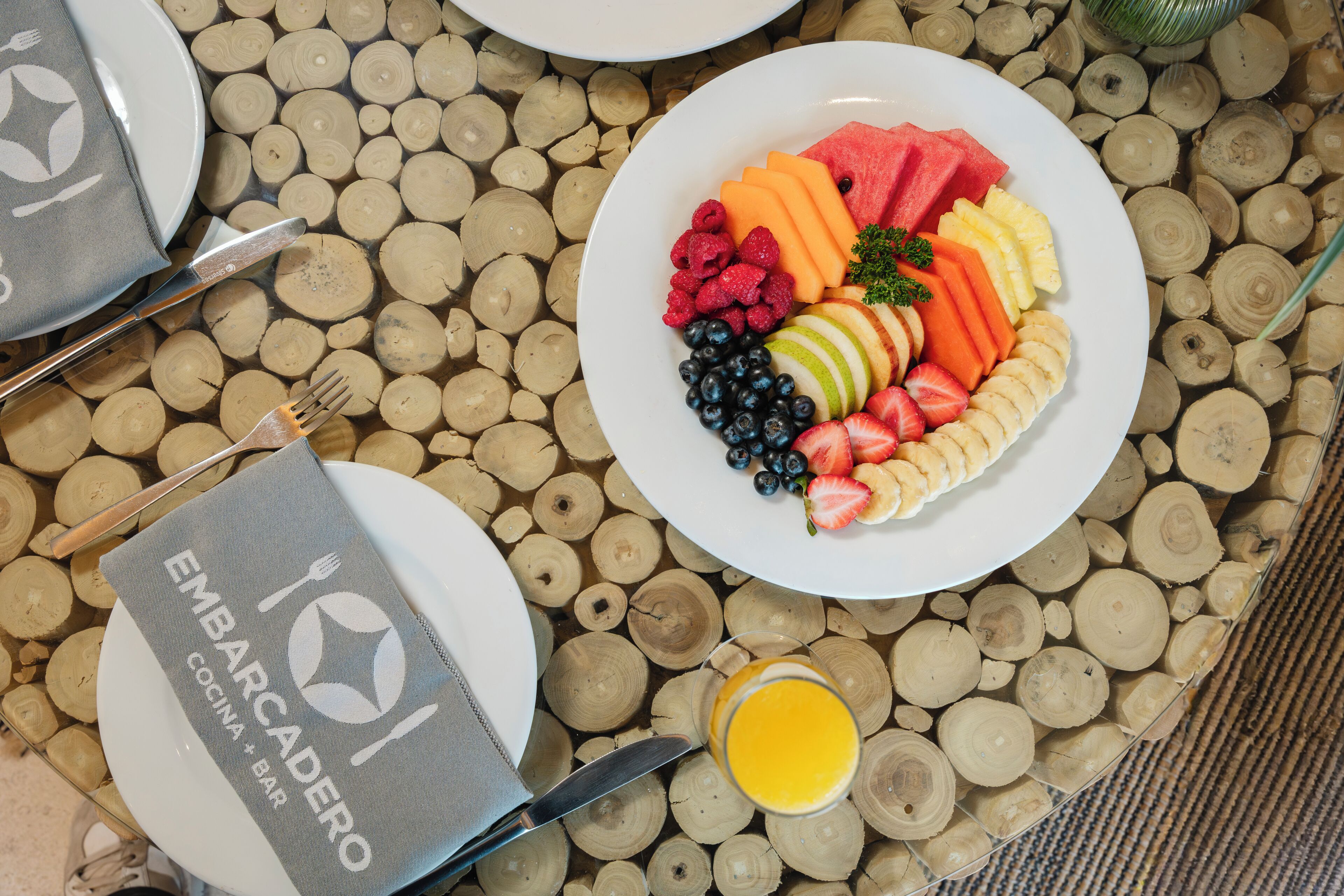Colazione con un piatto di frutta, un bicchiere di succo d'arancia e un tovagliolo con il marchio 'EMBARCADERO'.