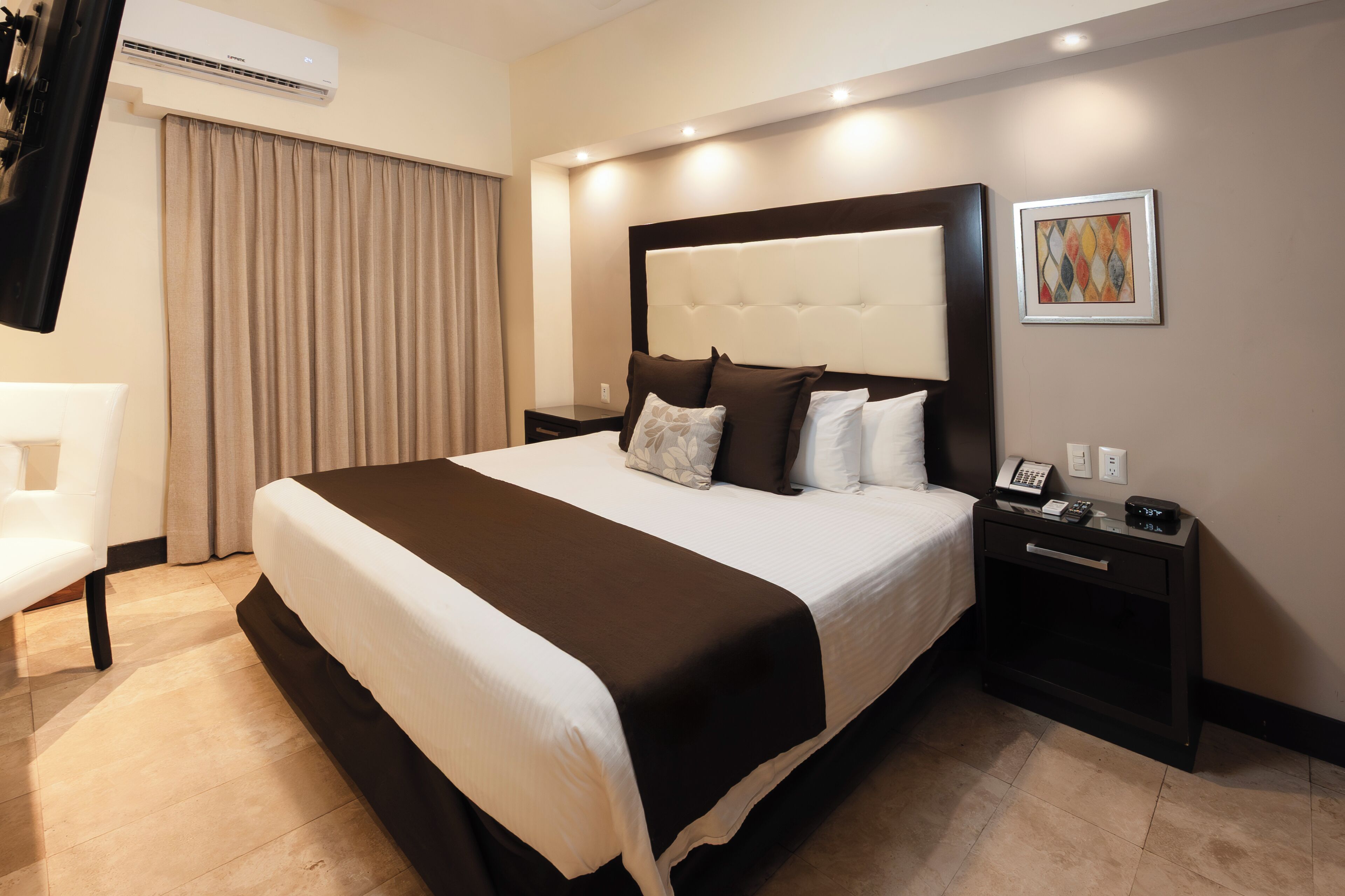 A hotel room with a large bed, a bedside table with a phone, a wall-mounted air conditioner, and a painting on the wall.