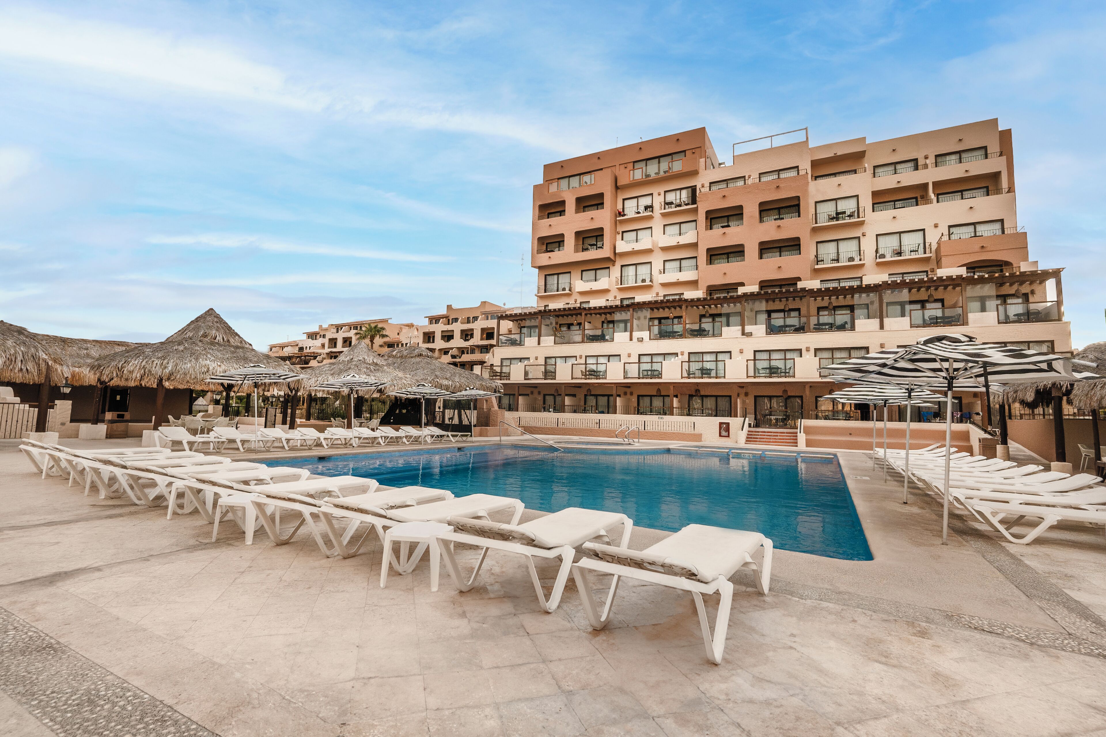 Una piscina con tumbonas, un edificio con varios balcones y sombrillas de paja.