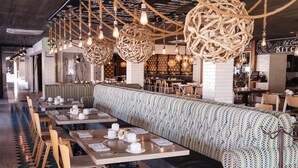 A restaurant with long communal tables, a patterned bench, and hanging wicker light fixtures.