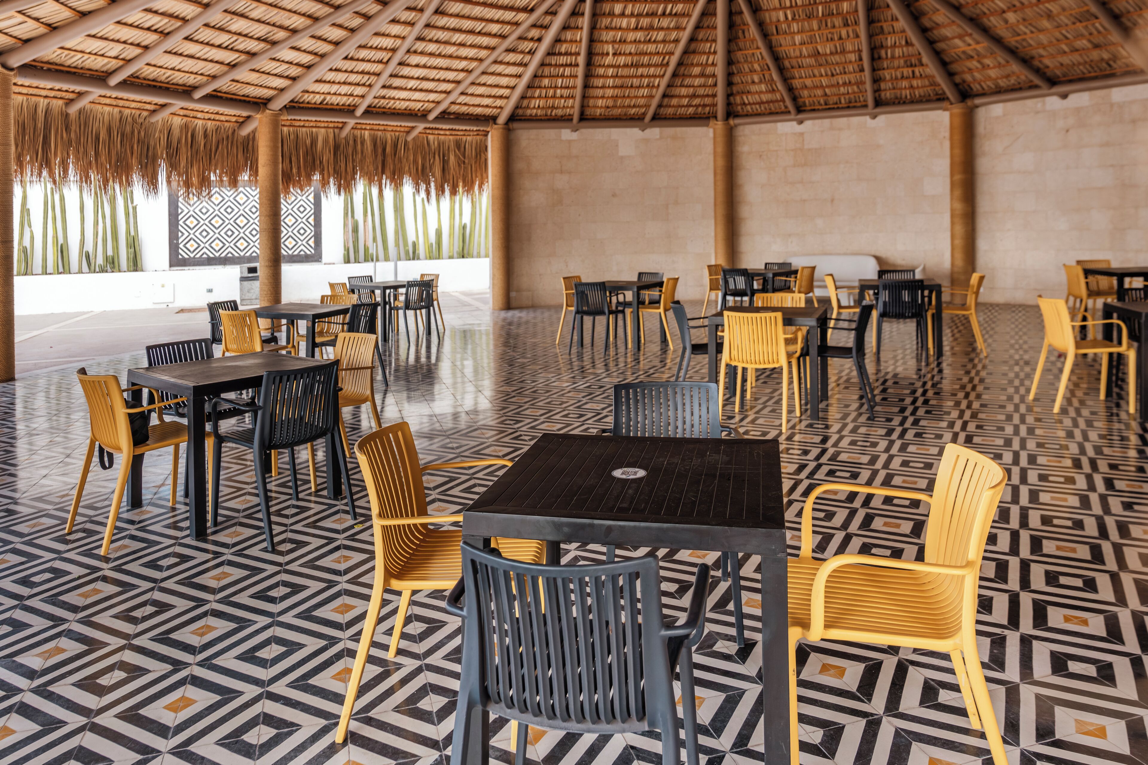Un’area pranzo all’aperto riparata, con tavoli e sedie, pavimento decorato e tetto in paglia.
