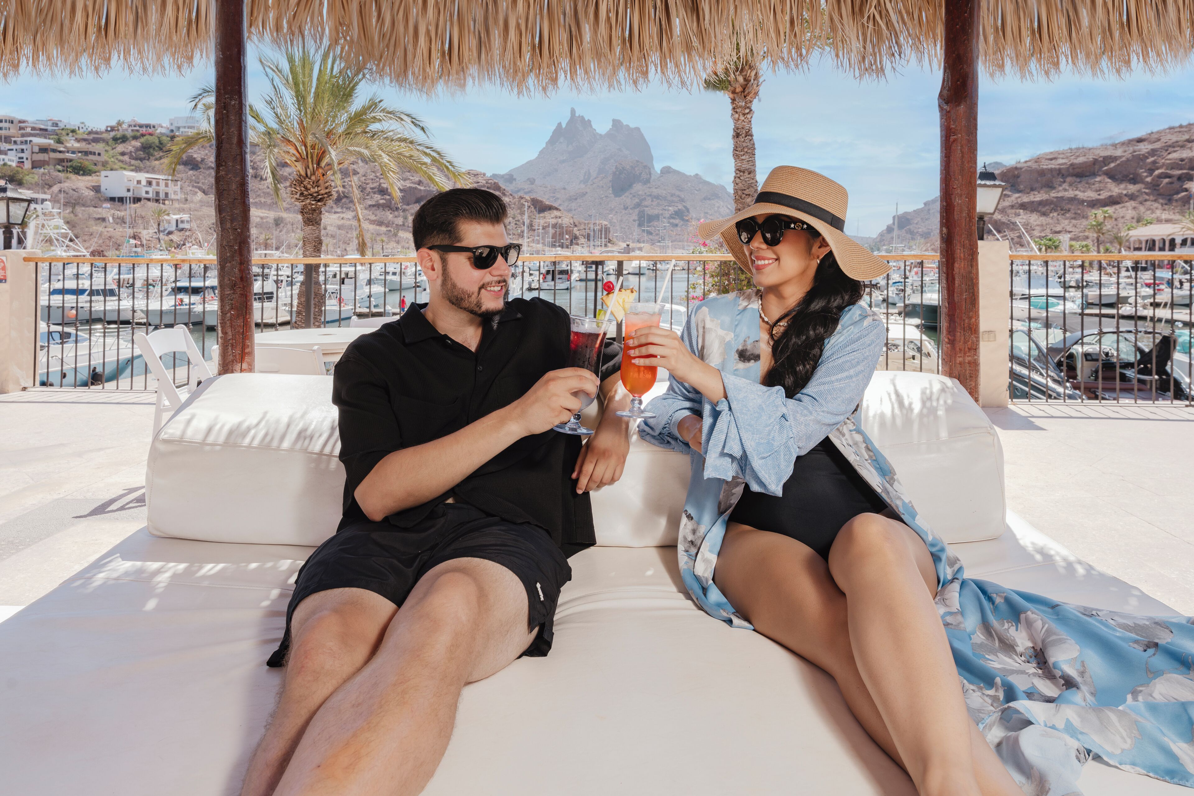 A couple sitting on a white outdoor sofa with a view of boats and mountains in the background.