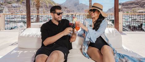 A couple sitting on a white outdoor sofa with a view of boats and mountains in the background.