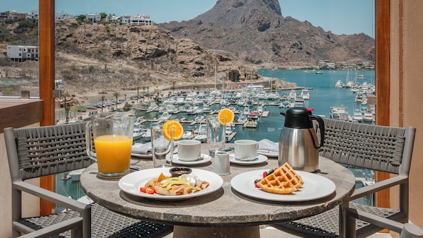 A table with breakfast items and a view of a marina with boats and a mountain.