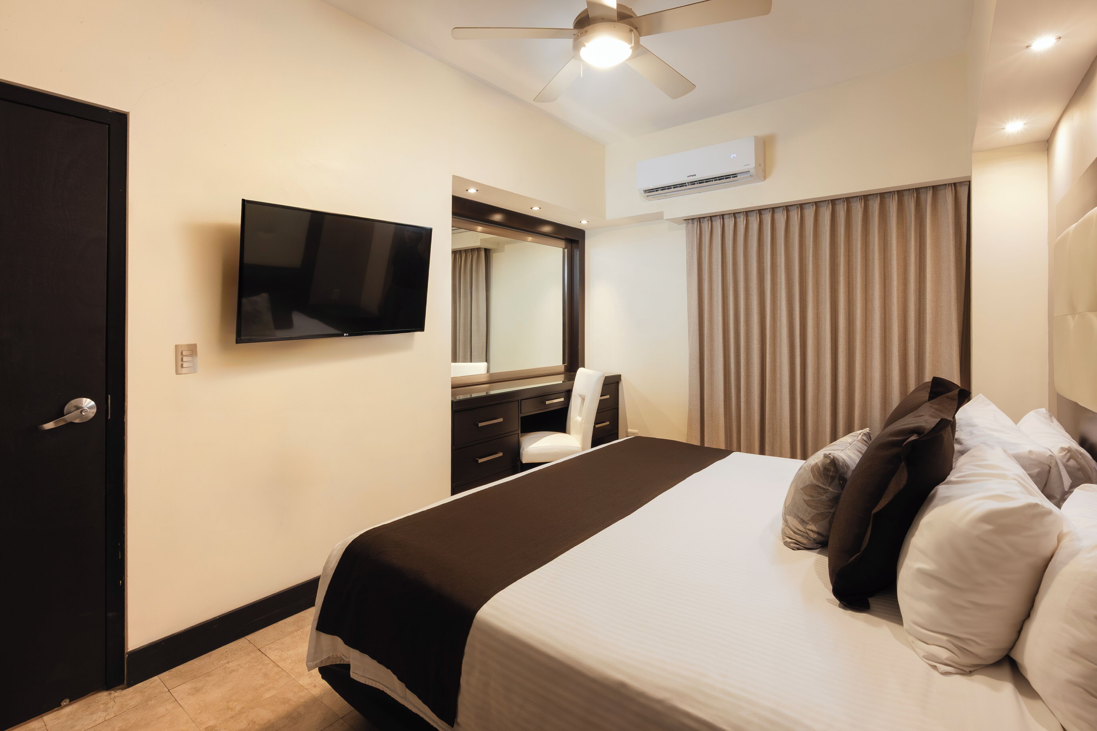 Ein Hotelzimmer mit Bett, Schreibtisch mit Stuhl, Fernseher, Deckenventilator und Klimaanlage.