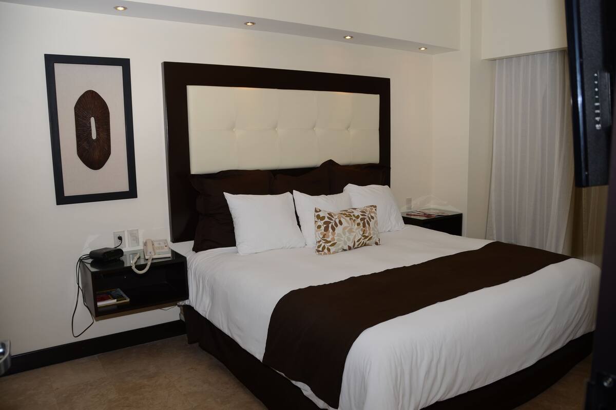 Una habitación de hotel con una cama grande, cabecera, mesita de noche con teléfono y un cuadro en la pared.