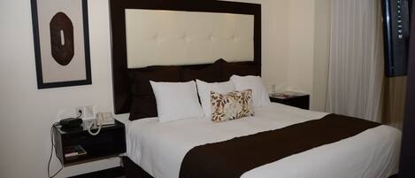Una habitación de hotel con una cama grande, cabecera, mesita de noche con teléfono y un cuadro en la pared.