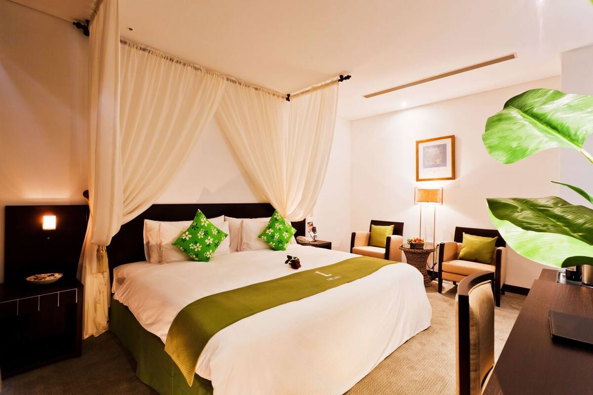 A hotel room with a bed, a nightstand, a chair, a desk, and a large plant.