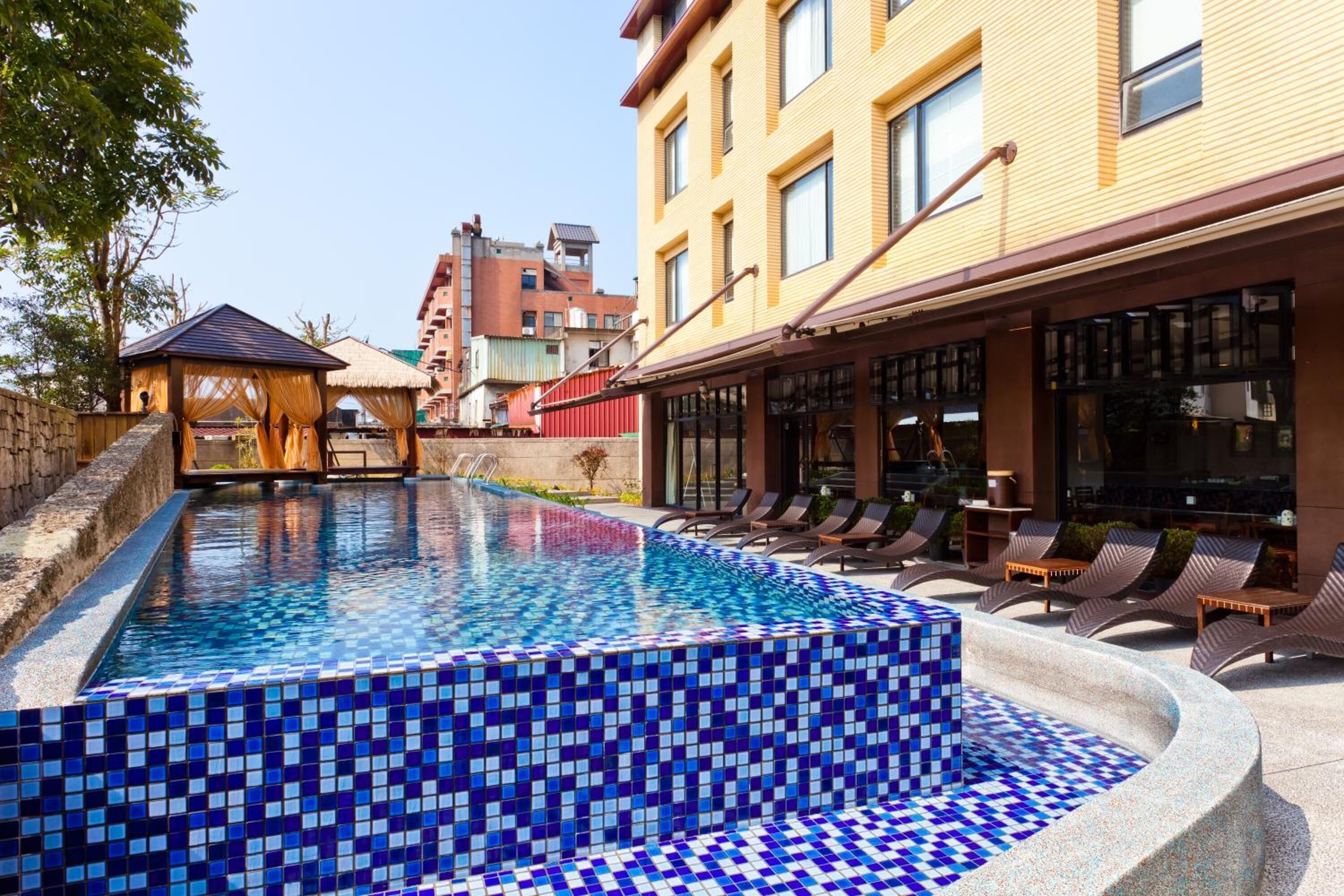 A swimming pool with blue mosaic tiles, lounge chairs, and a building with large windows.