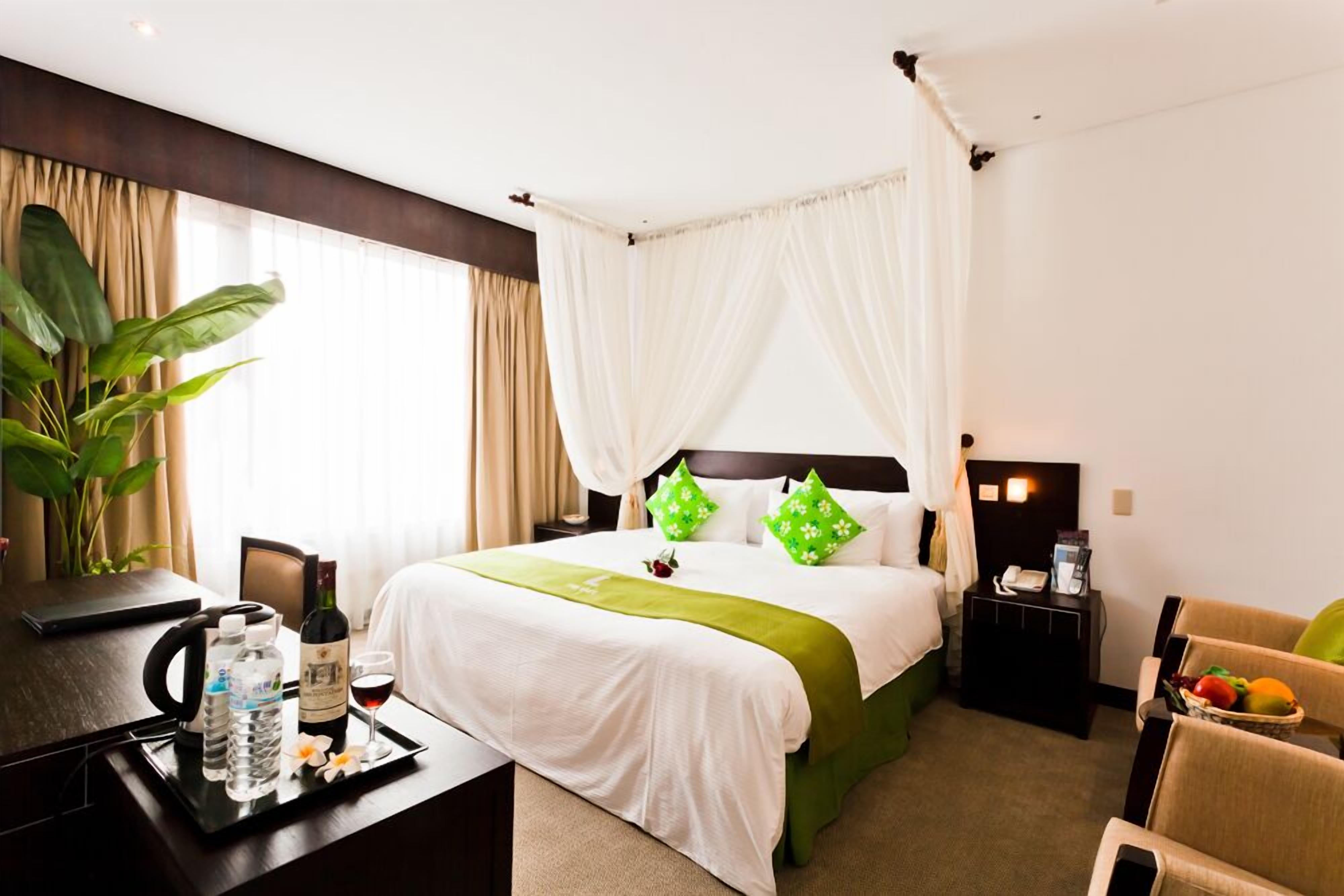 Habitación de hotel con cama, mesita de noche y escritorio con un atril de botellas de agua, una copa de vino y una botella.