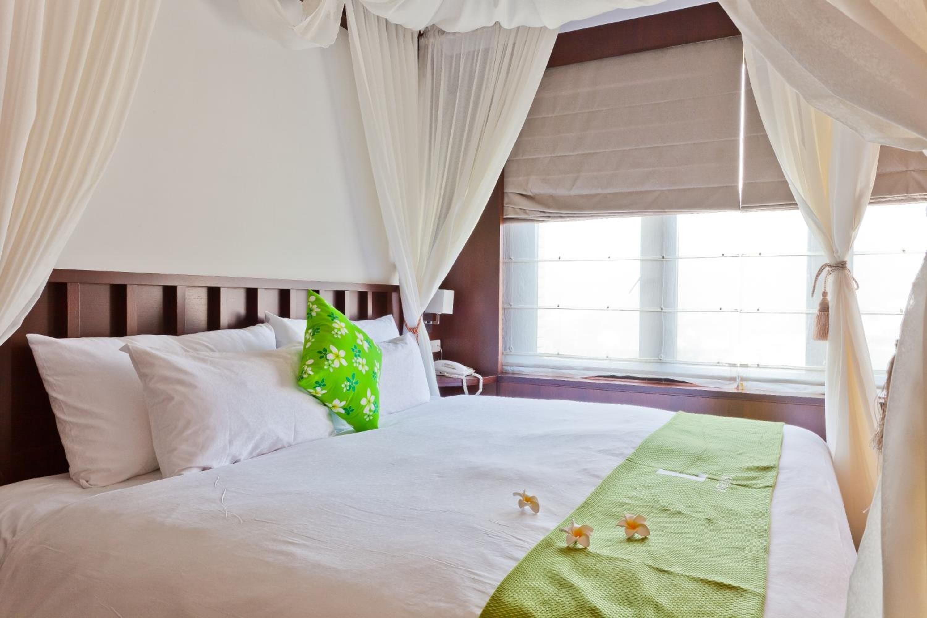 Een hotelkamer met een bed, witte gordijnen, een groen kussen en een raam met uitzicht.
