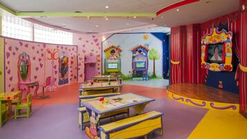 Ein helles Spielzimmer für Kinder mit Tischen, Stühlen und einer Bühne.