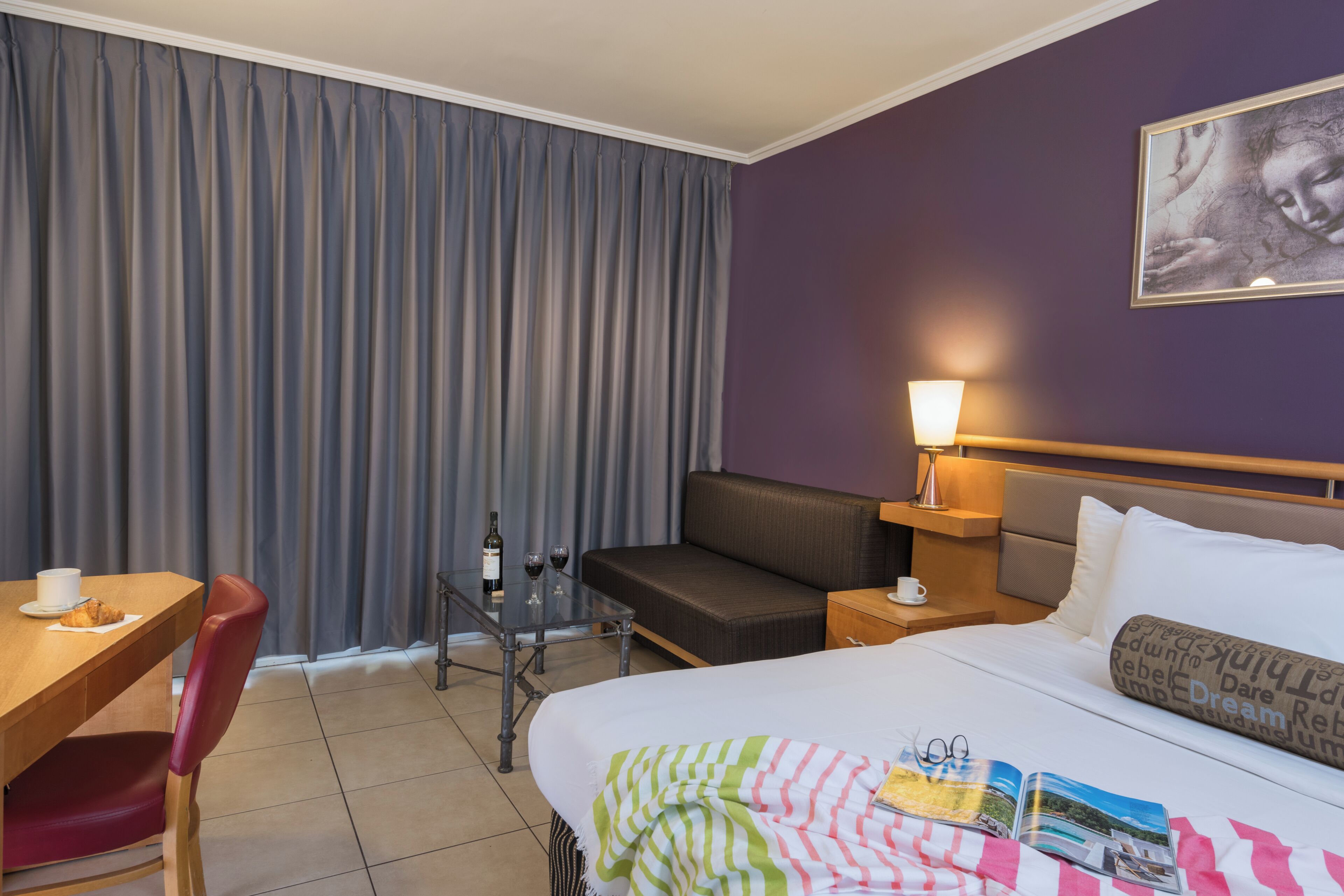 A hotel room with a bed, a desk with a chair, a sofa, a nightstand with a lamp, and a window with curtains.