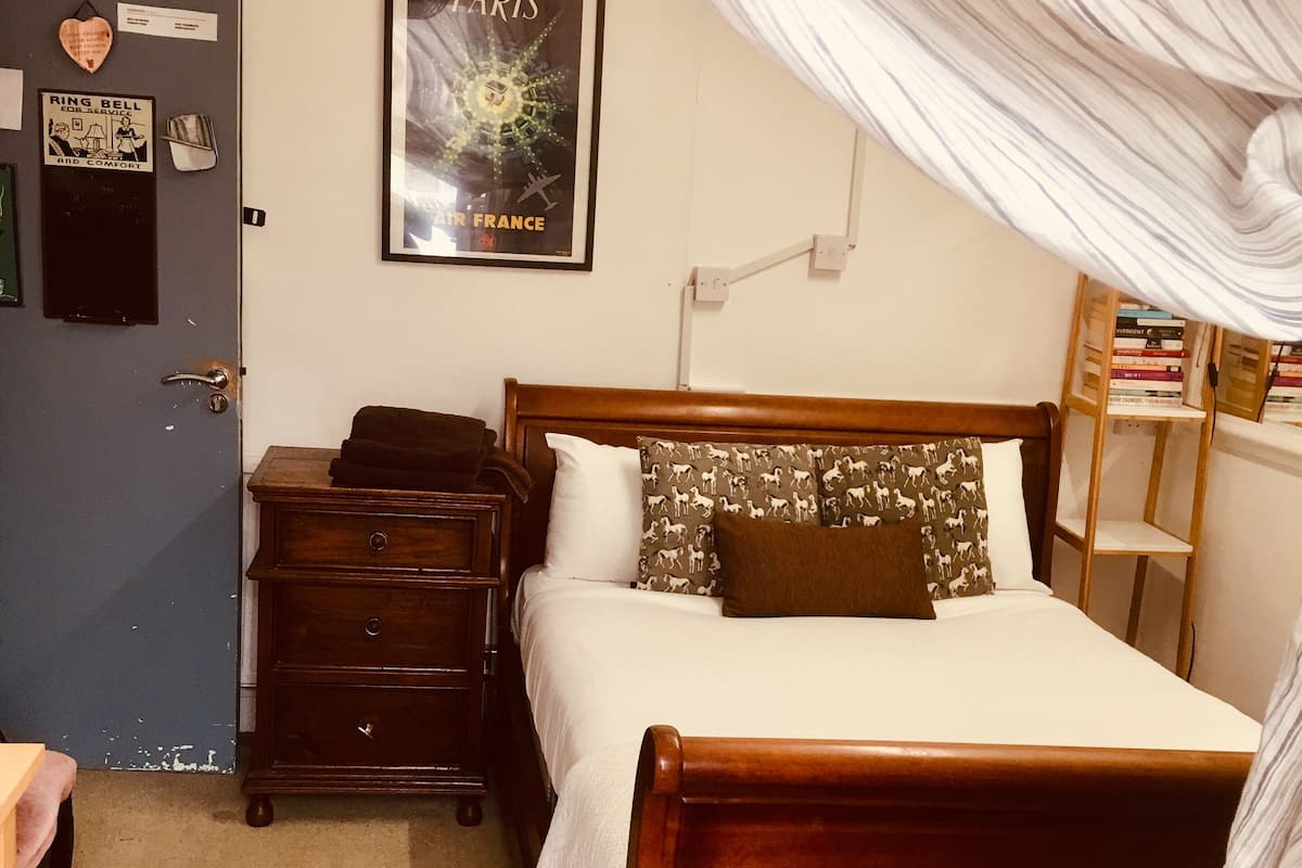 Quarto com cama de madeira, mesa de cabeceira, cômoda, pôster emoldurado e porta com placa.