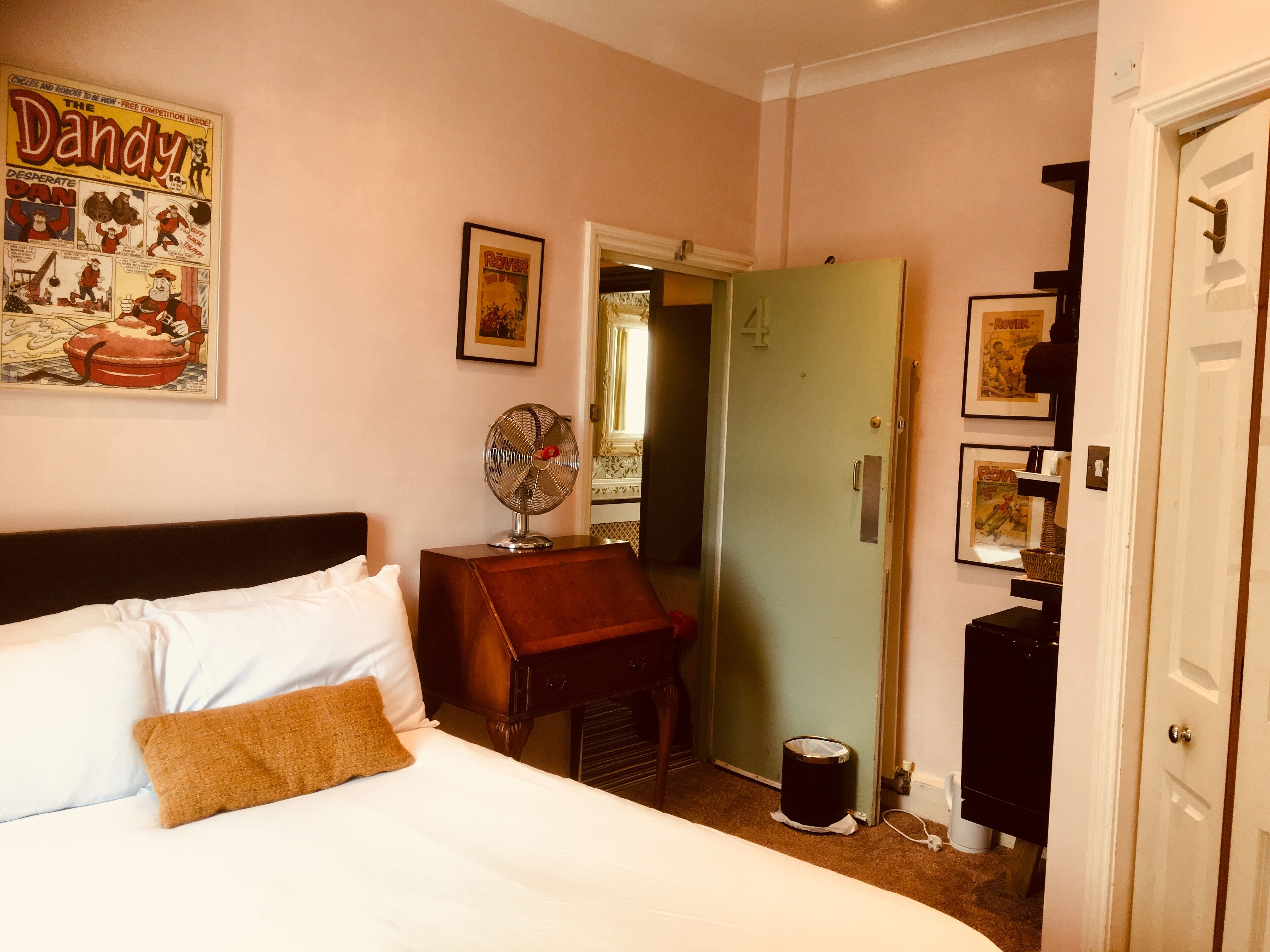 A hotel room with a bed, a small table with a fan, a door, and a poster on the wall.