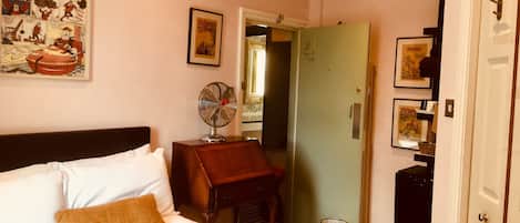 A hotel room with a bed, a small table with a fan, a door, and a poster on the wall.