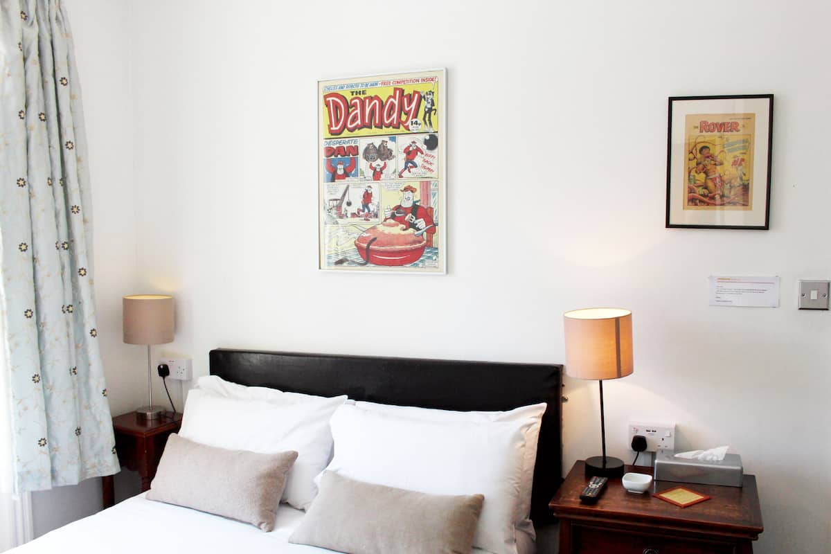 Quarto de hotel com uma cama, duas mesas de cabeceira, um criado-mudo, uma luminária, um cartaz emoldurado na parede e uma cortina.
