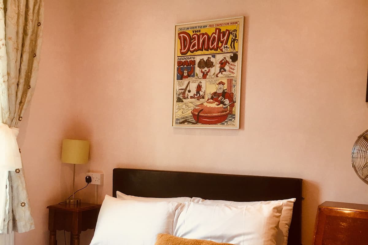 Quarto com cama, mesa de cabeceira, luminária, um pôster na parede e janela com cortinas.