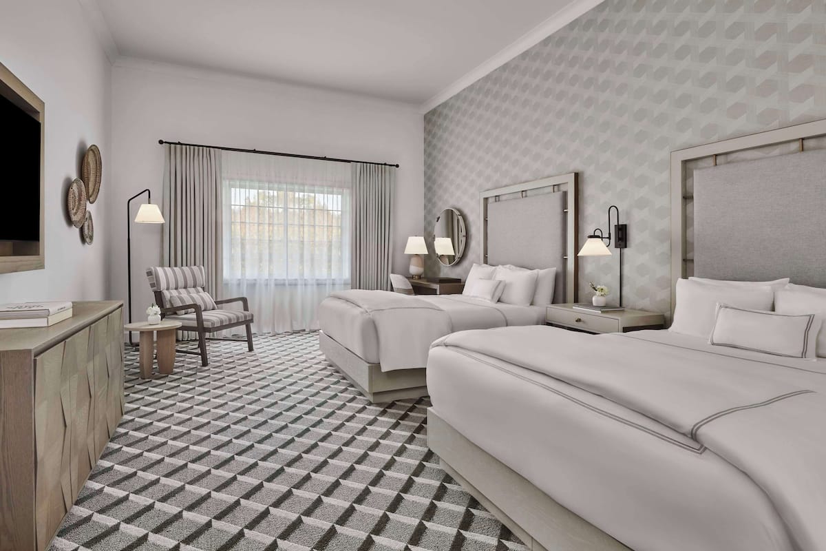 A hotel room with two beds, a chair, a desk, and a large window with curtains.