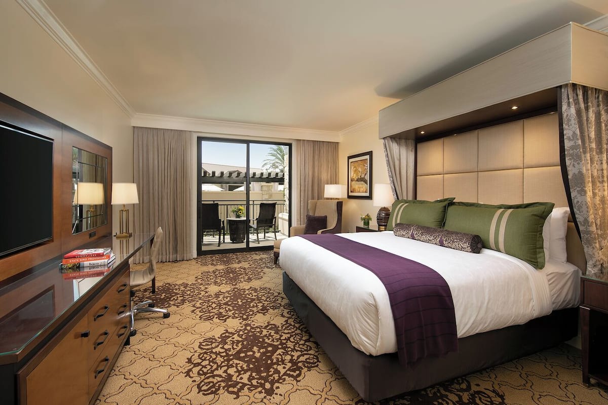 A hotel room with a large bed, a flat-screen TV, a desk with a lamp, and a view of the outdoors through a sliding door.