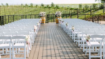 A wooden deck with white chairs arranged in rows, flanked by large flower arrangements.
