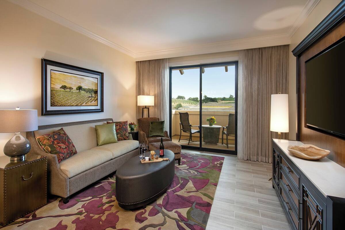 A hotel room with a sofa, armchair, and ottoman, a dining table with chairs, a TV, and a balcony with a view.