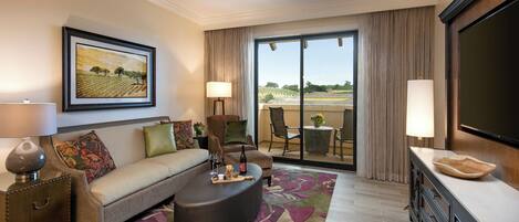 A hotel room with a sofa, armchair, and ottoman, a dining table with chairs, a TV, and a balcony with a view.