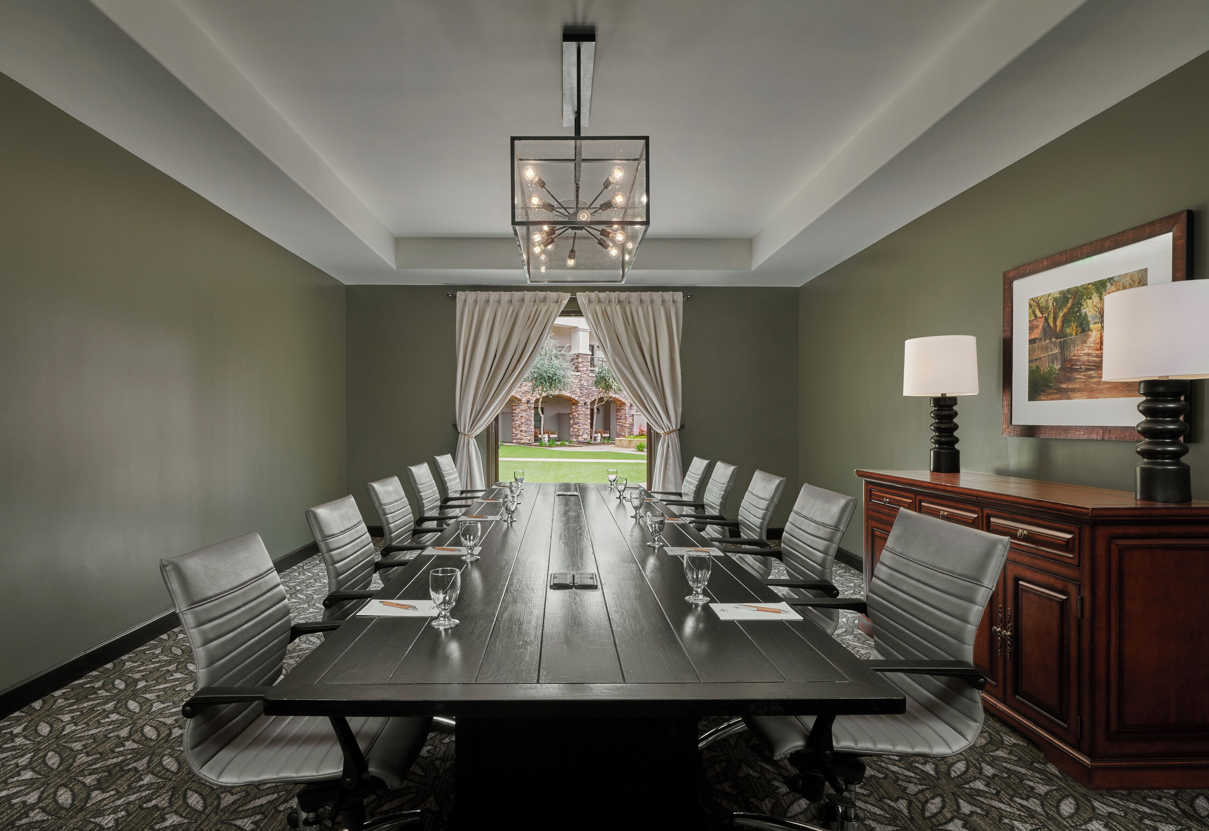A conference room with a long table, chairs, a chandelier, a desk with a lamp, and a framed picture on the wall.