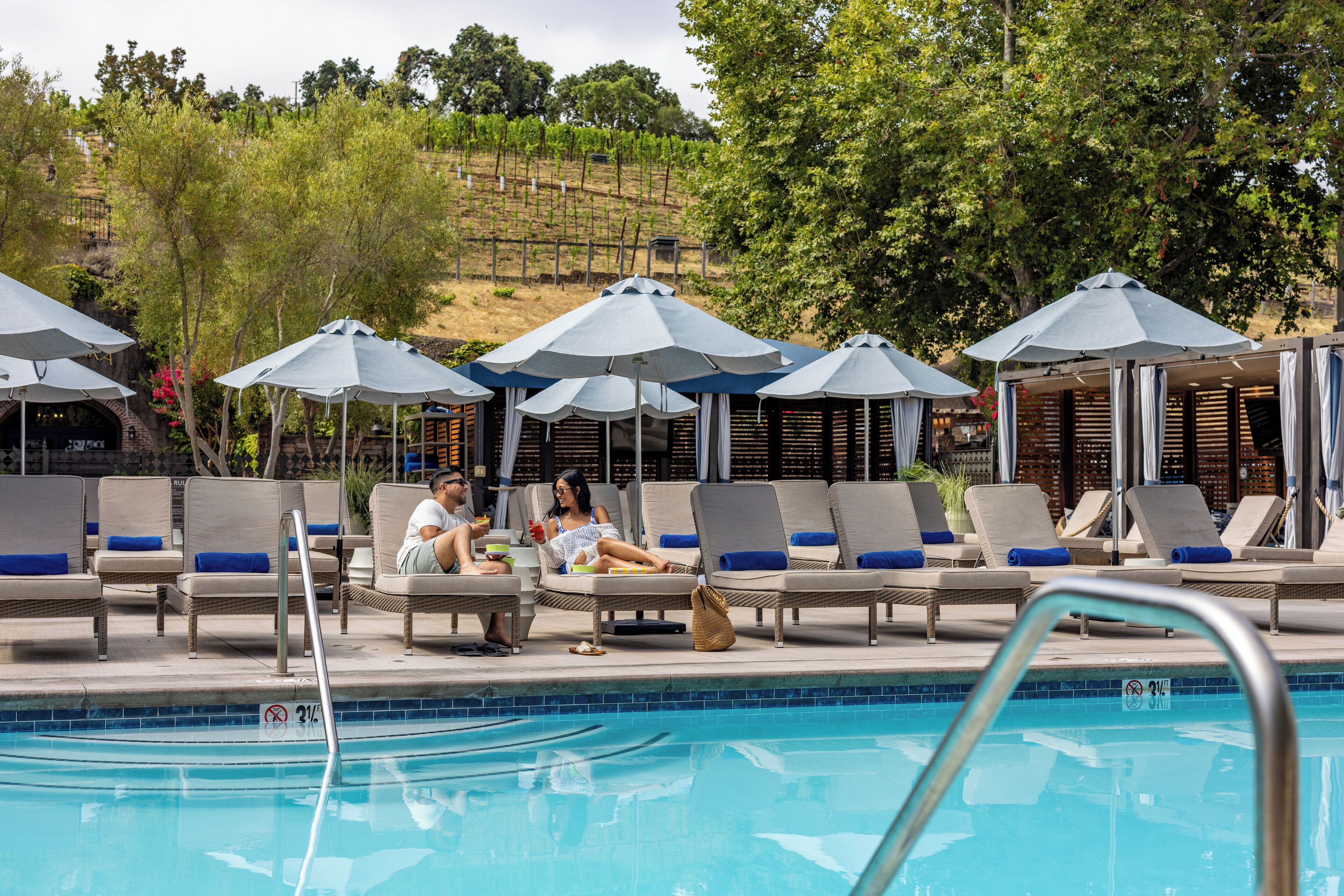 A poolside area with lounge chairs and umbrellas, a pool, and a hillside in the background.