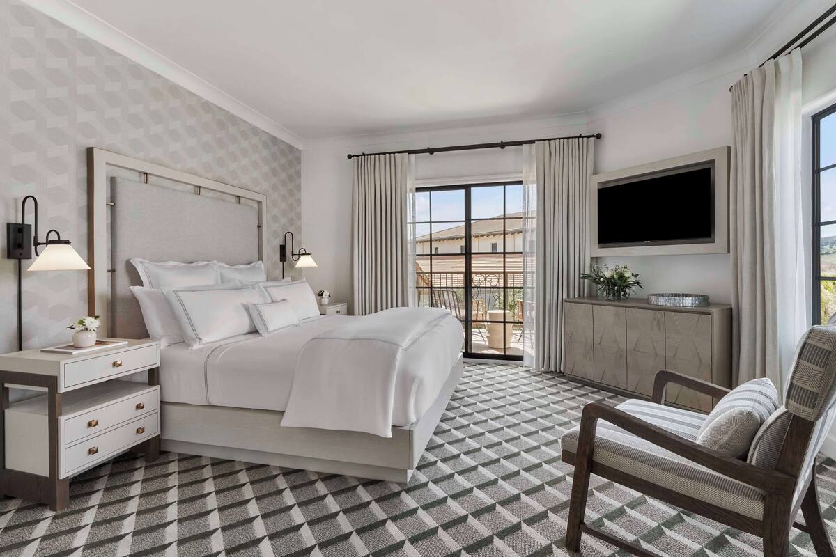 A hotel room with a bed, a TV mounted on the wall, a desk with drawers, a chair, and a patterned floor.