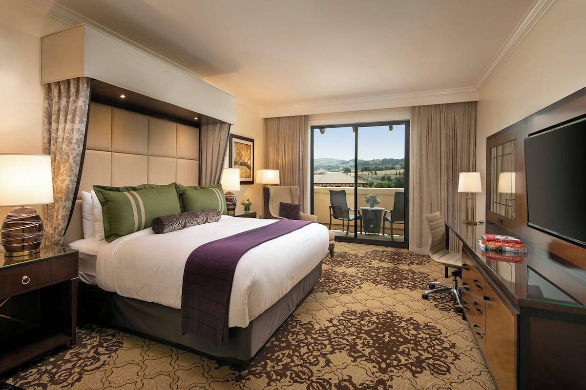 A hotel room with a large bed, a TV, a balcony with a view, and a desk with a lamp.