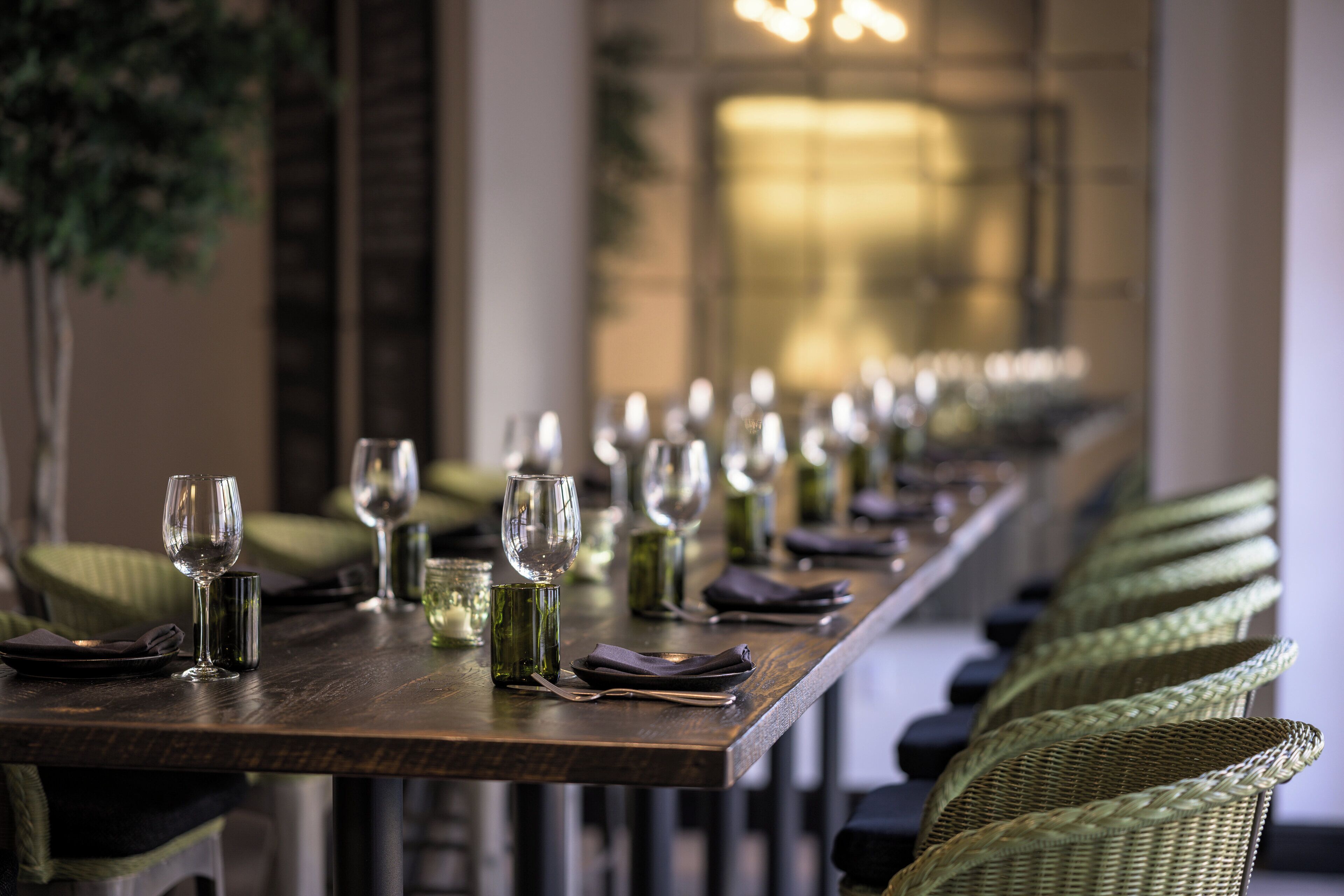 Une longue table à manger, dressée avec de nombreux verres à vin, des assiettes et des couverts, et entourée de chaises en osier.