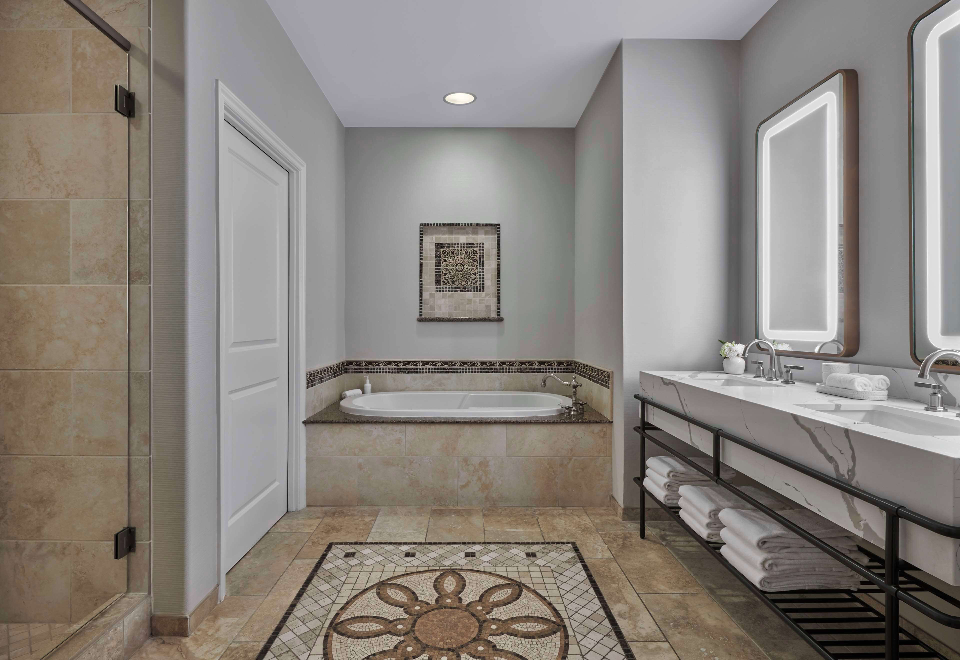 A modern bathroom with a bathtub, two sinks, a large mirror, and a patterned rug.